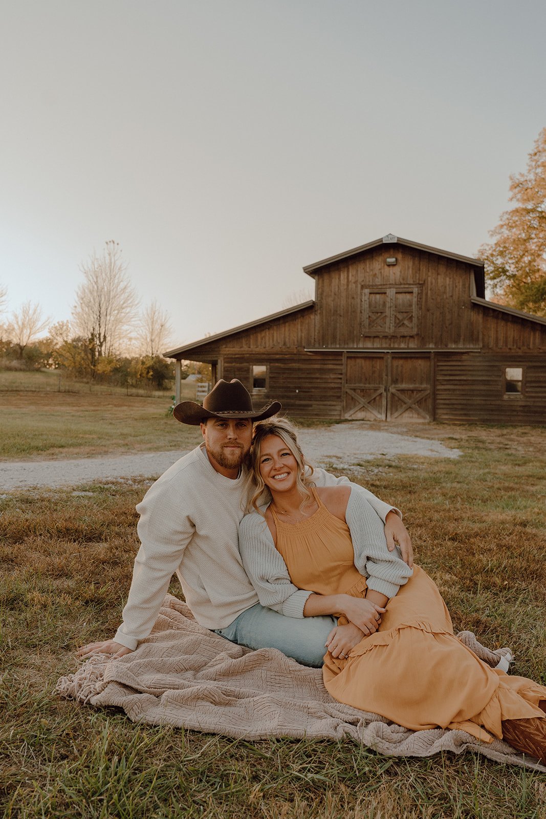 Liz and her husband sitting on a blanket in front of a weathered barn in autumn